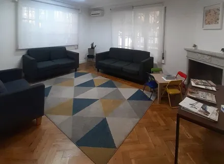 La sala de espera vista a lo ancho: dos sofás, la alfombra de triángulos, las ventanas a la calle y la chimenea.